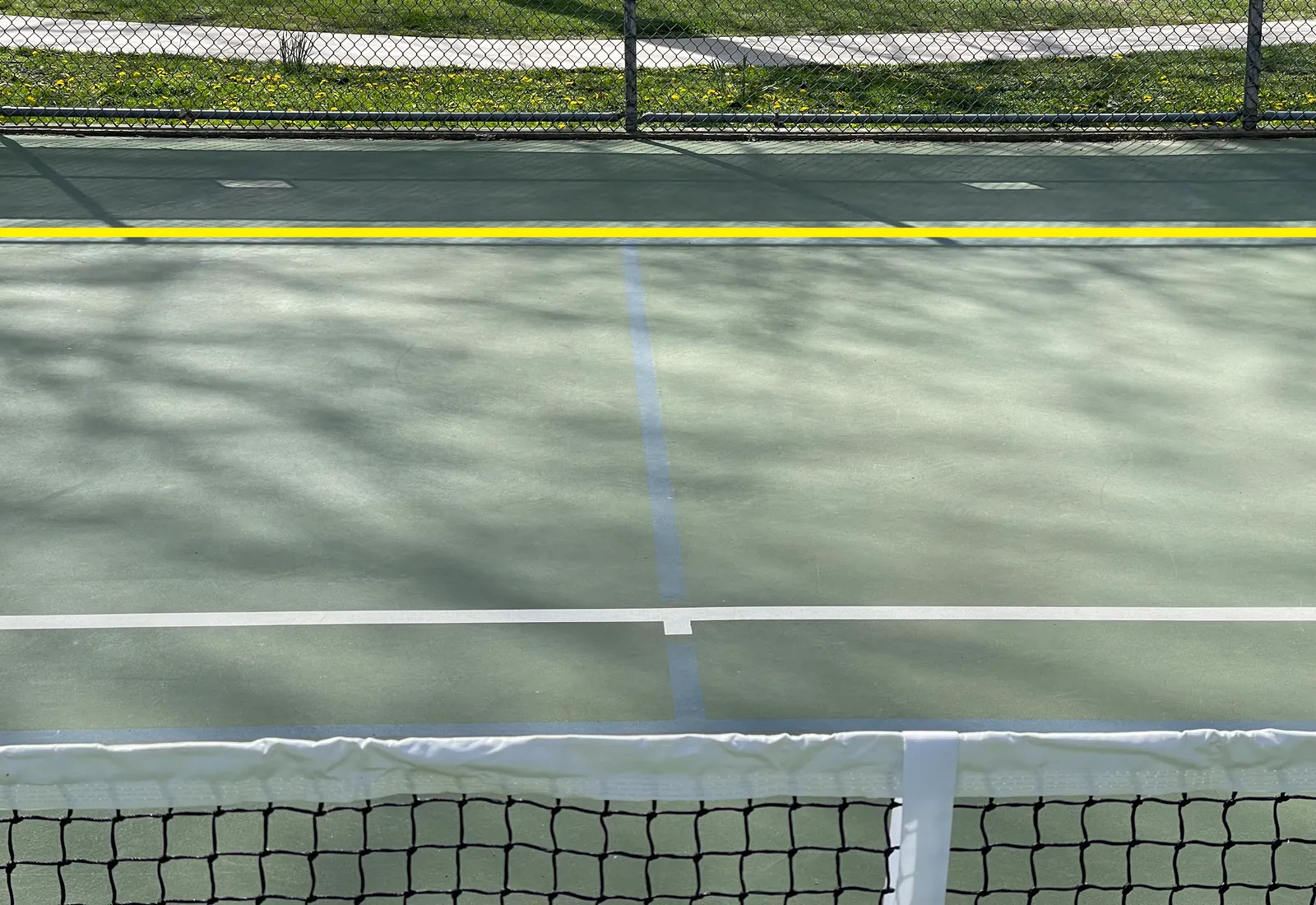 The same photo, edited: a bright yellow mark drawn on the image shows where the hidden pickleball baseline is, at the place where the lit court meets the shadow of the fence.
